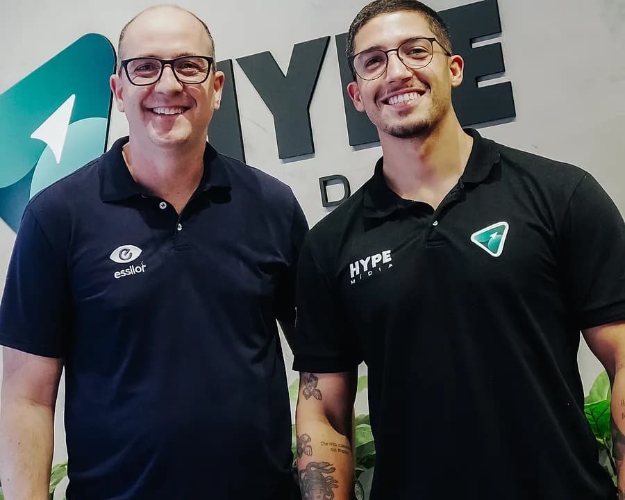 Dois homens lado a lado diante da marca da Hype Mídia aplicada na parede do escritório